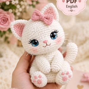 Pode incluir: Um brinquedo de gato em crochê branco com olhos azuis, cílios longos, interior das orelhas rosa e um laço rosa na cabeça. O gato tem almofadas rosas nas patas e um pequeno nariz rosa. O texto no canto superior direito diz: Crochet Pattern PDF English.