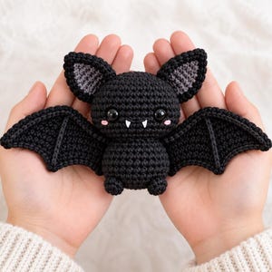 May include: A small, black crocheted bat plush toy featuring large, glossy black eyes, two small white fangs, and pink stitched cheeks. The bat has grey inner ears and wide, spread wings, crafted with a detailed yarn texture throughout its body.