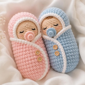 May include: Two crocheted baby dolls wrapped in swaddles, one in light pink and one in light blue. Each doll has a beige face, closed eyes with eyelashes, and a small pacifier matching its swaddle color. Two wooden buttons decorate each swaddle.