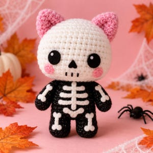 May include: A crocheted plush toy featuring a white skull head with pink ears and cheeks, large black eyes, and a black body decorated with white yarn bones. The figure has a rounded head and a compact body, showcasing detailed stitching throughout.