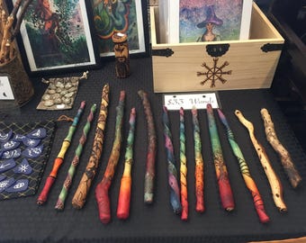 May include: A collection of handcrafted wands with colorful painted designs and natural wood textures. Some wands have spiral patterns and others feature intricate details. The display includes artwork and a wooden box with a symbol.