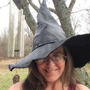 May include: A woman wearing a large black witch hat with a wide brim. She is smiling and looking at the camera. The hat is made of leather and has a pointed crown.