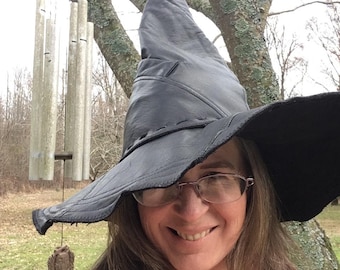 May include: A woman wearing a large black witch hat with a wide brim. She is smiling and looking at the camera. The hat is made of leather and has a pointed crown.