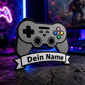 Könnte beinhalten: Ein graues Gaming-Controller-Schild mit einem schwarzen Steuerkreuz, zwei schwarzen Analogsticks und vier farbigen Aktionstasten in Pink, Blau, Gelb und Grün. Unter dem Controller befindet sich ein weißes Banner mit dem schwarzen Text Dein Name.