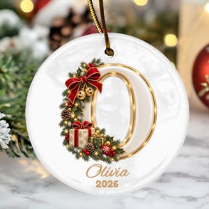 May include: A round white ceramic ornament featuring a large gold-trimmed letter O decorated with a festive wreath of pine branches, red bows, small bells, and gift boxes. The name Olivia and the year 2026 are printed in gold script below.