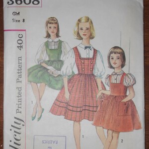 Può includere: Modello di abito per bambina, Simplicity Printed Pattern 40c, taglia 8. Il modello mostra tre bambine che indossano diverse versioni dell'abito. L'abito è a quadri rossi e bianchi con una camicetta bianca e un colletto alla Peter Pan. Il modello è da utilizzare con i tessuti Crownley's.
