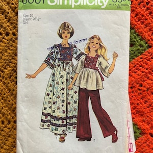 May include: Vintage Simplicity sewing pattern number 6001 for a girl's dress and pants. The pattern is for size 10 with a 28 1/2 inch bust. The pattern shows two girls wearing the outfits. The girl on the left is wearing a floral print dress with a white bodice and a brown belt. The girl on the right is wearing a white blouse with a brown peasant top and brown bell bottom pants.