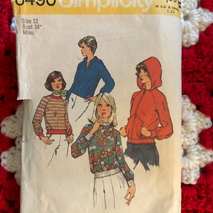 May include: A vintage Simplicity sewing pattern for a women's top. The pattern is for size 12, bust 34 inches. The pattern shows three different styles of tops, including a striped top, a floral top, and a solid top with a zipper. The pattern is labeled "6490 Simplicity" and "$1.00".
