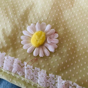 May include: A yellow fabric with a white daisy flower applique. The fabric has a small white polka dot pattern. The daisy has a yellow center and white petals. The fabric has a white lace trim.