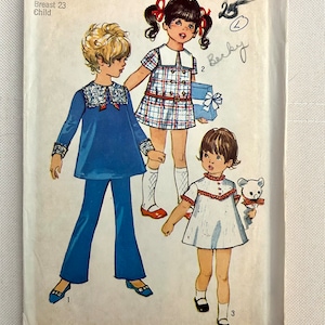 May include: Vintage Simplicity sewing pattern number 8567 for children's clothing. The pattern includes three different outfits: a blue dress with a white collar and cuffs, a plaid dress with a white collar and a belt, and a white dress with a red trim. The pattern is for sizes 4, with a 23-inch bust.