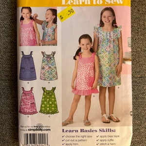 Könnte beinhalten: Simplicity Nähmuster Nummer 2241 für Mädchenkleider. Das Muster enthält Anleitungen zum Nähen von vier verschiedenen Kleidungsstücken, darunter ein rosa Kleid mit weißen Punkten, ein blaues Kleid mit weißen Punkten, ein grünes Kleid mit weißen Punkten und ein rosa Kleid mit weißen Blumen. Das Muster ist für die Größen K5, 7, 8, 10, 12 und 14. Das Muster enthält auch Anleitungen für grundlegende Nähtechniken, wie z. B. die Auswahl der richtigen Größe, das Zuschneiden eines Musters, das Anbringen von Besatz, das Anbringen eines Jochs, das Anbringen von Schrägband, das Anbringen eines Volants und das Nähen eines Saums.