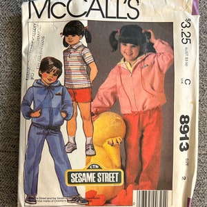May include: Sewing pattern for a Sesame Street doll outfit. The pattern is for size 2 and includes a pink jacket with a hood, red pants, a blue jacket with a hood, and blue pants. The pattern is from McCall's and has the number 8913.