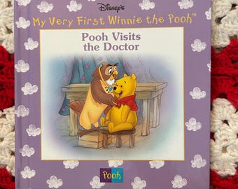 Puede incluir: Un libro infantil titulado "My Very First Winnie the Pooh: Pooh Visits the Doctor". La portada es morada con ilustraciones de nubes blancas. El libro presenta una ilustración de Winnie the Pooh y Búho. El logo de Pooh está en la parte inferior.