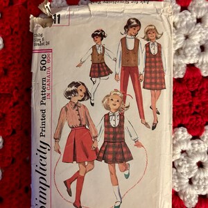 Peut inclure: Un patron imprimé Simplicity vintage pour enfants, avec des illustrations de filles portant des tenues telles qu'une robe à carreaux et un gilet avec un pantalon. Le patron est étiqueté "Child Size 6, Breast 24". Le prix est indiqué comme 50c au Canada.