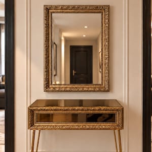 May include: A rectangular wall mirror with an ornate gold frame positioned above a matching console table. The console table features a dark mirrored top and front panel, an intricate gold frame, and four slender gold hairpin legs.