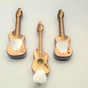 May include: Three wooden guitar-shaped bottle openers. The openers feature a light wood body with a darker wood neck and headstock. Each has a silver pick-shaped opener and a small brass screw. The guitars are approximately 5 inches long.