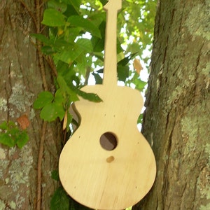 May include: A wooden guitar-shaped birdhouse is mounted on a tree trunk. The birdhouse is painted a light brown color and has a small hole for birds to enter.