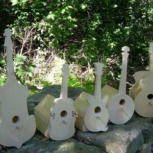 May include: Six wooden birdhouses shaped like different musical instruments, including a guitar, banjo, and violin. The birdhouses are all painted white and are sitting on a rock.