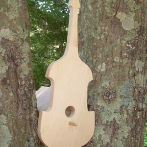 May include: A wooden birdhouse shaped like a bass guitar. The birdhouse is painted a light brown color and has a small hole for birds to enter. The birdhouse is mounted on a tree.