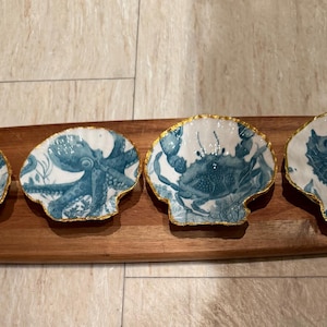 May include: Four white ceramic shell-shaped dishes with gold-painted edges, each featuring a blue illustration of a sea creature: a sea turtle, an octopus, a crab, and a seahorse. The dishes are arranged in a row on a rectangular wooden board.