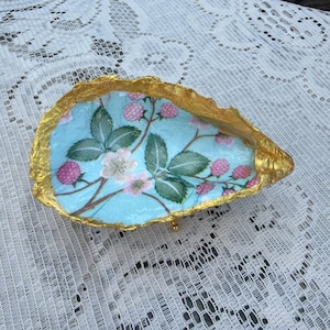 May include: A decorative oyster shell dish featuring a light blue interior with a botanical print of pink raspberries and green leaves. The irregular, textured edges of the shell are finished with a metallic gold paint, creating a distinct contrast.