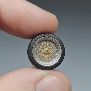 Könnte beinhalten: Ein Miniatur-Modellautorad mit einem schwarzen Gummireifen mit Michelin-Schriftzug und einer goldfarbenen Mehrspeichenfelge mit silbernem Außenrand. Das Rad verfügt über eine zentrale Nabenöffnung und detaillierte Speichen im gesamten Design.