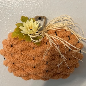 Pode incluir: Uma decoração de parede em forma de abóbora feita de fio laranja espesso e texturizado enrolado em torno de uma estrutura de arame. É adornada com uma flor de tecido amarelo pálido, folhas verdes e um feixe de ráfia natural no topo.
