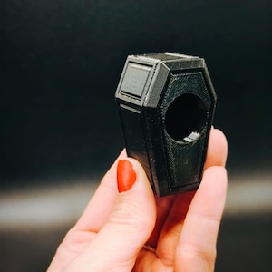 Könnte beinhalten: Ein kleines, schwarzes, 3D-gedrucktes Objekt in Sargform mit einer kreisförmigen Öffnung auf der Vorderseite. Die Oberfläche weist eine texturierte, geschichtete Struktur auf, die für additive Fertigung typisch ist, mit geometrischen Paneelen.