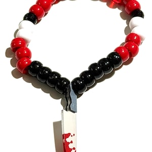 Op de afbeelding: Een kralenarmband met een cirkelvormige opstelling van witte, rode en zwarte plastic kralen. Een zilverkleurige metalen mesbedel hangt in het midden, met een rood geschilderd gedeelte op het lemmet dat lijkt op een bloedspatpatroon.