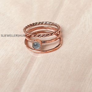 Puede incluir: Un conjunto de tres anillos de tono cobre apilados. El anillo superior presenta una textura de cuerda trenzada, mientras que el anillo central es liso y sostiene una pequeña piedra redonda de color azul claro. El anillo inferior es una banda simple y lisa de tono cobre. Texto: SLJEWELLERSHUB.