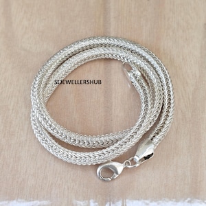 Puede incluir: Un collar de cadena de trigo en tono plateado con un diseño de eslabones tejidos y texturizados. El collar incluye un cierre de mosquetón en un extremo y está marcado con el texto SLJEWELLERSHUB. La cadena está enrollada en forma circular.