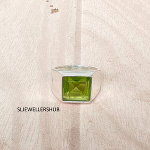 May include: A silver ring featuring a square-cut, light green peridot gemstone set in a polished metal band. The text SLJEWELLERSHUB is printed in black on the lower left side of the image, near the base of the ring.