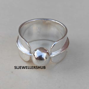May include: A polished silver ring featuring a wide band with a central spherical bead element. The design includes two curved side panels that frame the sphere. The text SLJELLERSHUB is printed in black on the lower left side of the image.