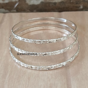 May include: Three silver-colored bangles stacked together, each featuring a repeating floral pattern along the band. The text SLJEWELLERSHUB is printed on the middle bangle. These circular bracelets have a polished, reflective metallic finish.