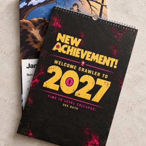May include: A wall calendar with a black cover featuring yellow text that reads NEW ACHIEVEMENT! WELCOME CRAWLER TO 2027 TIME TO LEVEL COLLAPSE: 365 DAYS. The design includes pink splatter accents and a spiral binding at the top edge of the paper.