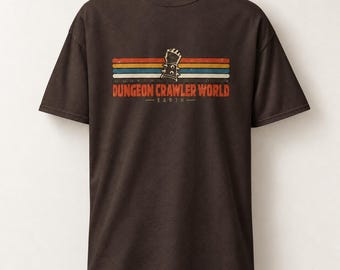Può includere: Una t-shirt a maniche corte marrone scuro con la grafica di un guanto chiodato centrato sopra strisce orizzontali rosse, arancioni e blu. Sotto le strisce, il testo "DUNGEON CRAWLER WORLD" in arancione, con la parola "EARTH" sottostante.