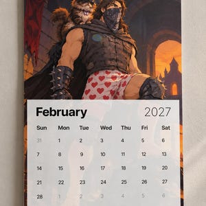 May include: A wall calendar for February 2027 featuring an illustration of a muscular figure wearing a dark vest, spiked arm guards, and heart-patterned boxers, with a fluffy cat wearing a crown and sunglasses perched on their shoulder.