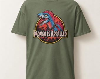 Può includere: Una t-shirt a maniche corte color verde oliva con una grafica circolare sul petto. La grafica raffigura un dinosauro piumato blu e rosso con un collare all'interno di un cerchio con bordo giallo e la scritta MONGO IS APPALLED in bianco.