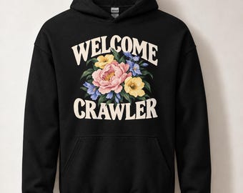 Può includere: Una felpa con cappuccio nera caratterizzata da una stampa grafica di un bouquet floreale con fiori rosa, gialli e blu. Il testo WELCOME è stampato in un carattere color crema arcuato sopra i fiori, e CRAWLER è scritto sotto.