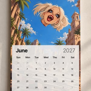 May include: A wall calendar for June 2027 featuring a spiral binding at the top. The illustration depicts a floating head with blonde hair, blue eyes, and visible fangs against a bright blue sky, palm trees, and stone ruins. The calendar grid is white.