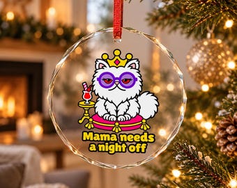 Può includere: Un ornamento in vetro circolare trasparente appeso a un nastro rosso. Presenta la grafica di un gatto bianco soffice con una corona dorata e occhiali viola, seduto su un cuscino rosa con un cocktail e il testo Mama needs a night off.