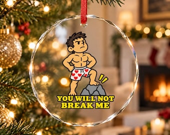 Può includere: Un ornamento in vetro trasparente, circolare e sfaccettato, appeso a un nastro rosso. Presenta un'illustrazione di un personaggio muscoloso in boxer bianchi con cuori, in piedi su una roccia con il testo, YOU WILL NOT BREAK ME.
