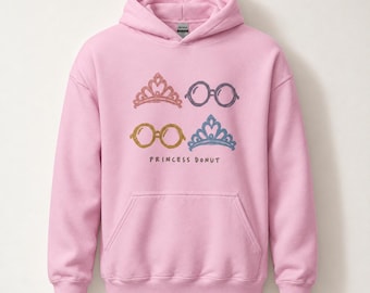 Può includere: Una felpa con cappuccio rosa chiaro con una grafica frontale raffigurante due diademi e due paia di occhiali rotondi in colori tenui. Sotto la grafica, il testo PRINCESS DONUT è stampato con un carattere semplice e scuro.