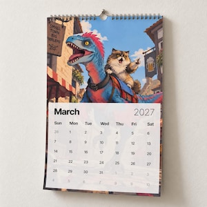 May include: A wall calendar for March 2027 featuring an illustration of a blue and red dinosaur with a fluffy cat riding on its back. The scene includes a town background with a sign reading Small Town Big Problems and a beer mug icon sign.