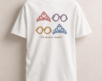 Può includere: Una t-shirt bianca a maniche corte con una stampa grafica di due diademi e due paia di occhiali rotondi in rosso, viola, oro e blu. Il testo "PRINCESS DONUT" è stampato in lettere marroni sotto le icone illustrate sul davanti.