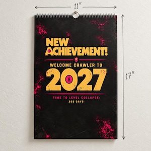 May include: A black wall calendar with pink and yellow text reading NEW ACHIEVEMENT! WELCOME CRAWLER TO 2027 TIME TO LEVEL COLLAPSE: 365 DAYS. The calendar features a pink eye graphic inside the zero and measures 11 inches by 17 inches.