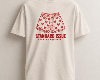Può includere: Una t-shirt a maniche corte color crema con una grafica rossa raffigurante dei boxer coperti di piccoli cuori. Sotto la grafica, il testo STANDARD ISSUE CRAWLER EQUIPMENT è stampato in grassetto rosso in lettere maiuscole.