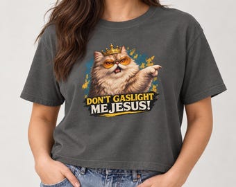 Può includere: Una t-shirt grigio antracite con la grafica di un gatto soffice che indossa una corona d'oro e occhiali con lenti arancioni. Il gatto punta una zampa in avanti. Sotto il gatto, il testo recita DON'T GASLIGHT ME JESUS! in lettere gialle e bianche.