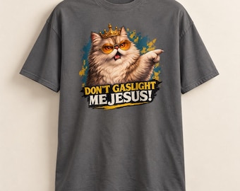 May include: A charcoal gray t-shirt featuring a graphic of a fluffy cat wearing a gold crown and orange-tinted glasses. The cat is pointing a paw forward. Below the cat, the text reads DON'T GASLIGHT ME JESUS! in bold yellow and white letters.