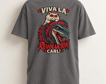 Può includere: Una t-shirt grigio antracite con una stampa grafica di un soffice gatto bianco che indossa una corona e occhiali da sole rossi, a cavallo di un dinosauro blu e rosso. Il testo VIVA LA REVOLUCION CARL! è stampato in rosso e bianco con stelle.